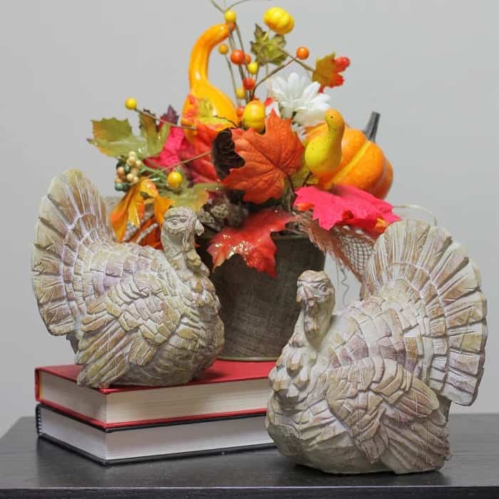two resin turkey decorations displayed with books and a bucket of fall leaves