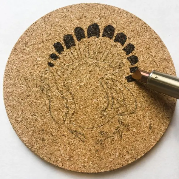 wood burning tool filling in small areas of a cork coaster with a stenciled turkey design