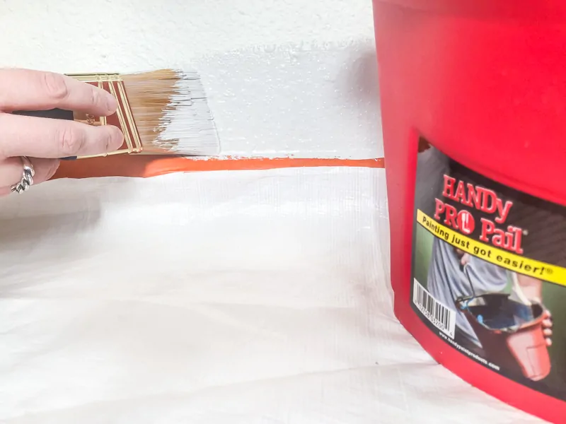 painting edge of baseboards above painter's tape with paint brush with HANDy PRO Pail in foreground