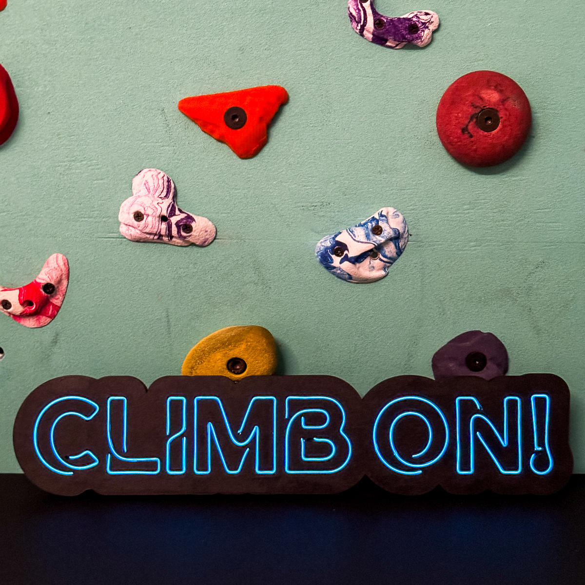 DIY LED neon sign that says "Climb on!" in front of a climbing wall